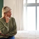 Senior woman sage-sweater, jeans sitting on bed in bedroom touching face facing window, cream cover Senior woman sage-sweater, jeans sitting on bed in bedroom touching face facing window, cream cover - PhotoDune Item for Sale