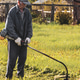 An elderly man in workwear mows grass and dandelions with an electric mower An elderly man in workwear mows grass and dandelions with an electric mower - PhotoDune Item for Sale