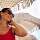 Smiling woman in red swimsuit wearing sunglasses, relaxing under a fringed beach umbrella Smiling woman in red swimsuit wearing sunglasses, relaxing under a fringed beach umbrella - PhotoDune Item for Sale