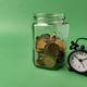 A retro style alarm clock in front of a glass jar filled with coins A retro style alarm clock in front of a glass jar filled with coins - PhotoDune Item for Sale