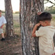 Young boy with short hair hides behind tree, covering face, while two other children play Young boy with short hair hides behind tree, covering face, while two other children play - PhotoDune Item for Sale