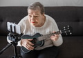 Elderly woman 70+ sits on the couch and learns to play the ukulele at home using video tutorial Elderly woman 70+ sits on the couch and learns to play the ukulele at home using video tutorial - PhotoDune Item for Sale