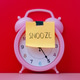 A pink alarm clock with a yellow sticky note taped over its face with the handwritten word SNOOZE. A pink alarm clock with a yellow sticky note taped over its face with the handwritten word SNOOZE. - PhotoDune Item for Sale