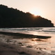 Golden hour sun peaking behind lush hill casting warm light on ocean waves Golden hour sun peaking behind lush hill casting warm light on ocean waves - PhotoDune Item for Sale