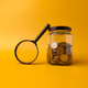 Clear glass jar filled with coins and a magnifying glass on a yellow background Clear glass jar filled with coins and a magnifying glass on a yellow background - PhotoDune Item for Sale