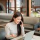 Smiling asian woman talking on phone open one palm over laptop and book while sits at table in cafe. Smiling asian woman talking on phone open one palm over laptop and book while sits at table in cafe. - PhotoDune Item for Sale