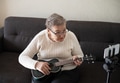 Elderly woman 70+ sits on the couch and learns to play the ukulele at home using video tutorial Elderly woman 70+ sits on the couch and learns to play the ukulele at home using video tutorial - PhotoDune Item for Sale