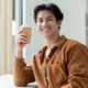 A handsome, positive Asian man sits at a table with a coffee cup in his hand, smiling at the camera. A handsome, positive Asian man sits at a table with a coffee cup in his hand, smiling at the camera. - PhotoDune Item for Sale