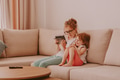 Modern interior in cozy home. Kids using technologies. Video call chat. Sisters sitting on couch Modern interior in cozy home. Kids using technologies. Video call chat. Sisters sitting on couch - PhotoDune Item for Sale