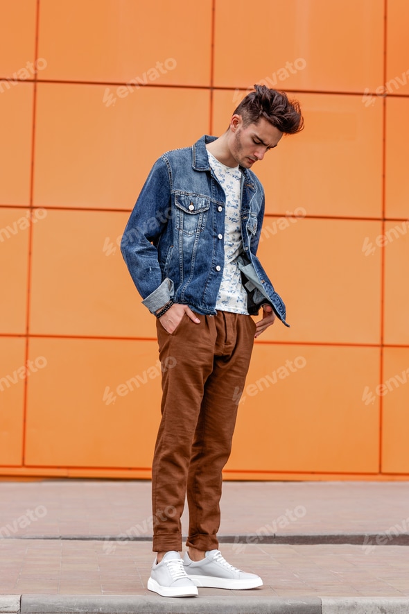 Young guy dressed in casual clothes brown trousers and jeans jacket poses in the street on the Stock Photo by leikapro Young guy dressed in casual clothes brown trousers and jeans jacket poses in the street on the Stock Photo by leikapro