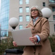 A woman in a brown coat works on her laptop outdoors in an urban area with modern buildings and lamp A woman in a brown coat works on her laptop outdoors in an urban area with modern buildings and lamp - PhotoDune Item for Sale