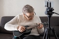 Elderly woman 70+ sits on the couch and learns to play the ukulele at home using video tutorial Elderly woman 70+ sits on the couch and learns to play the ukulele at home using video tutorial - PhotoDune Item for Sale