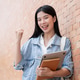Young asian woman student holding laptop and book raising a fist standing leaning against brick wall Young asian woman student holding laptop and book raising a fist standing leaning against brick wall - PhotoDune Item for Sale