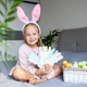 Young girl wearing bunny ears holding handmade paper crafts beside a basket of colorful eggs indoors Young girl wearing bunny ears holding handmade paper crafts beside a basket of colorful eggs indoors - PhotoDune Item for Sale