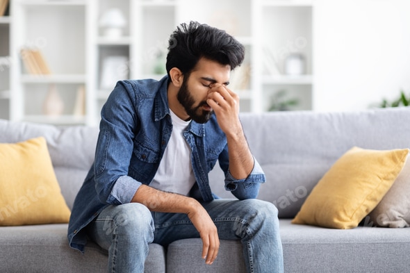 Seasonal Depression. Portrait Of Sad Indian Man Sitting On Couch At Home Stock Photo by Prostock-studio