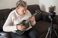 Elderly woman 70+ sits on the couch and learns to play the ukulele at home using video tutorial Elderly woman 70+ sits on the couch and learns to play the ukulele at home using video tutorial - PhotoDune Item for Sale