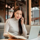 Pretty asian woman talking on phone as holding notebook aside laptop while sitting at table in cafe. Pretty asian woman talking on phone as holding notebook aside laptop while sitting at table in cafe. - PhotoDune Item for Sale