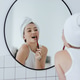 Blonde with a white towel on her head brushes her teeth in front of a mirror in a white bathroom. Blonde with a white towel on her head brushes her teeth in front of a mirror in a white bathroom. - PhotoDune Item for Sale