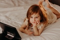 Toddler girl lying on bed in modern room, using laptop tablet pad gadget, looking at camera Toddler girl lying on bed in modern room, using laptop tablet pad gadget, looking at camera - PhotoDune Item for Sale
