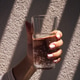 Hand holding a clear glass of sparkling water with bubbles, sunlight casting shadows Hand holding a clear glass of sparkling water with bubbles, sunlight casting shadows - PhotoDune Item for Sale