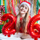 Smiling girl in Santa hat holding red number balloons at festive party Smiling girl in Santa hat holding red number balloons at festive party - PhotoDune Item for Sale