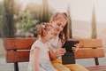 Two little girls sitting on bench with tablet pad and having video chat with friends Two little girls sitting on bench with tablet pad and having video chat with friends - PhotoDune Item for Sale