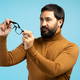 Bearded man holding eyeglasses and looking away on blue background Bearded man holding eyeglasses and looking away on blue background - PhotoDune Item for Sale