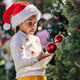 A joyful child wearing a Santa hat decorates a Christmas tree outdoors A joyful child wearing a Santa hat decorates a Christmas tree outdoors - PhotoDune Item for Sale