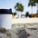 white paper cup of coffee is standing in the sand on the beach, in the back of a palm tree, sunny da white paper cup of coffee is standing in the sand on the beach, in the back of a palm tree, sunny da - PhotoDune Item for Sale