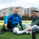 Two men sit on soccer field with ball and water bottle near city buildings on cloudy day Two men sit on soccer field with ball and water bottle near city buildings on cloudy day - PhotoDune Item for Sale