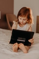 Toddler girl sitting on bed, using laptop tablet pad gadget, watching with smiling face Toddler girl sitting on bed, using laptop tablet pad gadget, watching with smiling face - PhotoDune Item for Sale