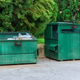 Two green dumpsters positioned beside concrete wall in parking area surrounded by trees on clear day Two green dumpsters positioned beside concrete wall in parking area surrounded by trees on clear day - PhotoDune Item for Sale