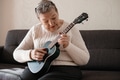 Elderly woman 70+ sits on the couch at home and plays the ukulele Elderly woman 70+ sits on the couch at home and plays the ukulele - PhotoDune Item for Sale