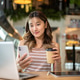 Pretty asian woman holding coffee looking at phone in front of a laptop on table in cafe or canteen. Pretty asian woman holding coffee looking at phone in front of a laptop on table in cafe or canteen. - PhotoDune Item for Sale