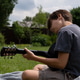 Young boy sitting outdoors on a blanket, focused on playing an acoustic guitar Young boy sitting outdoors on a blanket, focused on playing an acoustic guitar - PhotoDune Item for Sale