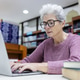 An elderly woman with gray hair and glasses works on a laptop in a library. An elderly woman with gray hair and glasses works on a laptop in a library. - PhotoDune Item for Sale