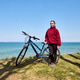 Young woman standing with blue bicycle on grassy coast under clear blue sky Young woman standing with blue bicycle on grassy coast under clear blue sky - PhotoDune Item for Sale