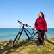 Young woman standing with bicycle on grassy cliff overlooking calm ocean under clear blue sky Young woman standing with bicycle on grassy cliff overlooking calm ocean under clear blue sky - PhotoDune Item for Sale