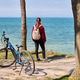 Young woman standing beside bicycle on sandy path overlooking calm blue ocean under clear sky Young woman standing beside bicycle on sandy path overlooking calm blue ocean under clear sky - PhotoDune Item for Sale