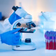 Scientist in Lab Coat and Blue Gloves Examining Slide Under Microscope with Blood Sample Test Tubes Scientist in Lab Coat and Blue Gloves Examining Slide Under Microscope with Blood Sample Test Tubes - PhotoDune Item for Sale