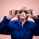 Older woman wearing blue coat holds oversized glasses in front of her face, with a pink wall backgro Older woman wearing blue coat holds oversized glasses in front of her face, with a pink wall backgro - PhotoDune Item for Sale