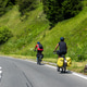 Two cyclists ride loaded bicycles winding mountain road green alpine hillside bright summer sun. Sce Two cyclists ride loaded bicycles winding mountain road green alpine hillside bright summer sun. Sce - PhotoDune Item for Sale