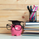 Piggy bank with glasses and graduation cap next to stacked notebooks and pens on a table Piggy bank with glasses and graduation cap next to stacked notebooks and pens on a table - PhotoDune Item for Sale