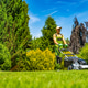 Man mows grass in a garden with trees and blue sky in the background Man mows grass in a garden with trees and blue sky in the background - PhotoDune Item for Sale