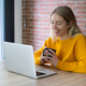 Smiling young woman in a yellow sweater holding a coffee mug and looking at a laptop during a cozy h Smiling young woman in a yellow sweater holding a coffee mug and looking at a laptop during a cozy h - PhotoDune Item for Sale