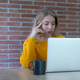 A thoughtful young woman in a yellow sweater works on a laptop at a table with a coffee mug against A thoughtful young woman in a yellow sweater works on a laptop at a table with a coffee mug against - PhotoDune Item for Sale