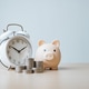 Piggy bank with stacked coins beside a white alarm clock on a wooden table, symbolizing payment remi Piggy bank with stacked coins beside a white alarm clock on a wooden table, symbolizing payment remi - PhotoDune Item for Sale