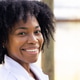 Mid adult African woman smiling at camera outdoors near wooden post, wearing white coat Mid adult African woman smiling at camera outdoors near wooden post, wearing white coat - PhotoDune Item for Sale