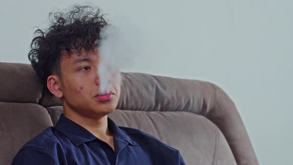 Young Asian man sitting on sofa exhaling thick vapor from electronic cigarette or vape device, Stock Footage Young Asian man sitting on sofa exhaling thick vapor from electronic cigarette or vape device, Stock Footage